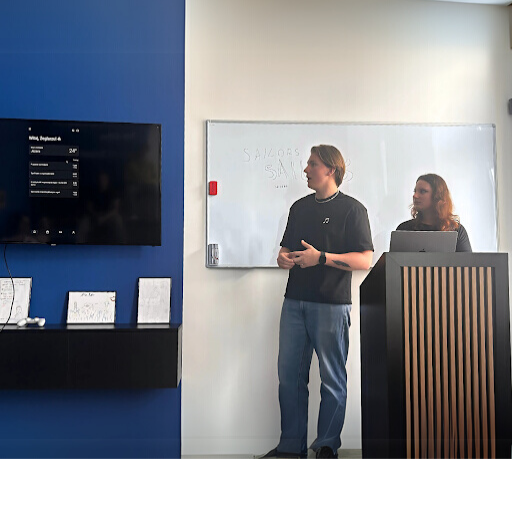A young man and a young woman are giving a presentation in a modern office setting. The man is standing and speaking in front of a whiteboard, while the woman is behind a lectern with a laptop.